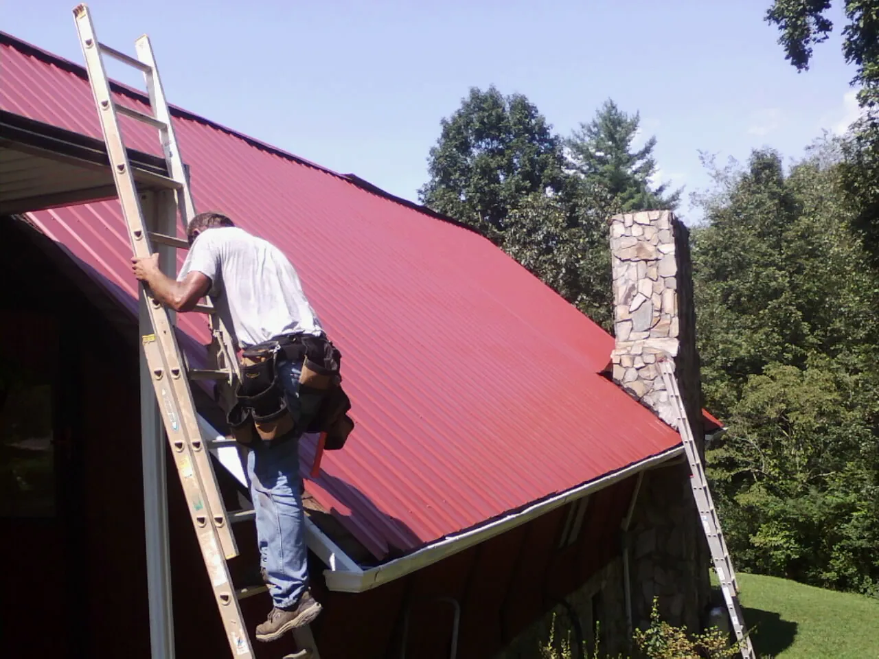 Roofer on a ladder beside a red metal roof and stone chimney — Soffit & Fascia Repair by Roofing Brothers in Tooele